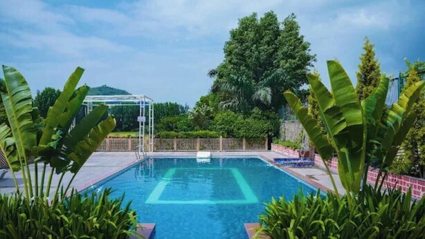A swimming pool with a clear blue center, surrounded by lush greenery and a pergola structure. - Abha Farms And Resort (Udaipur)