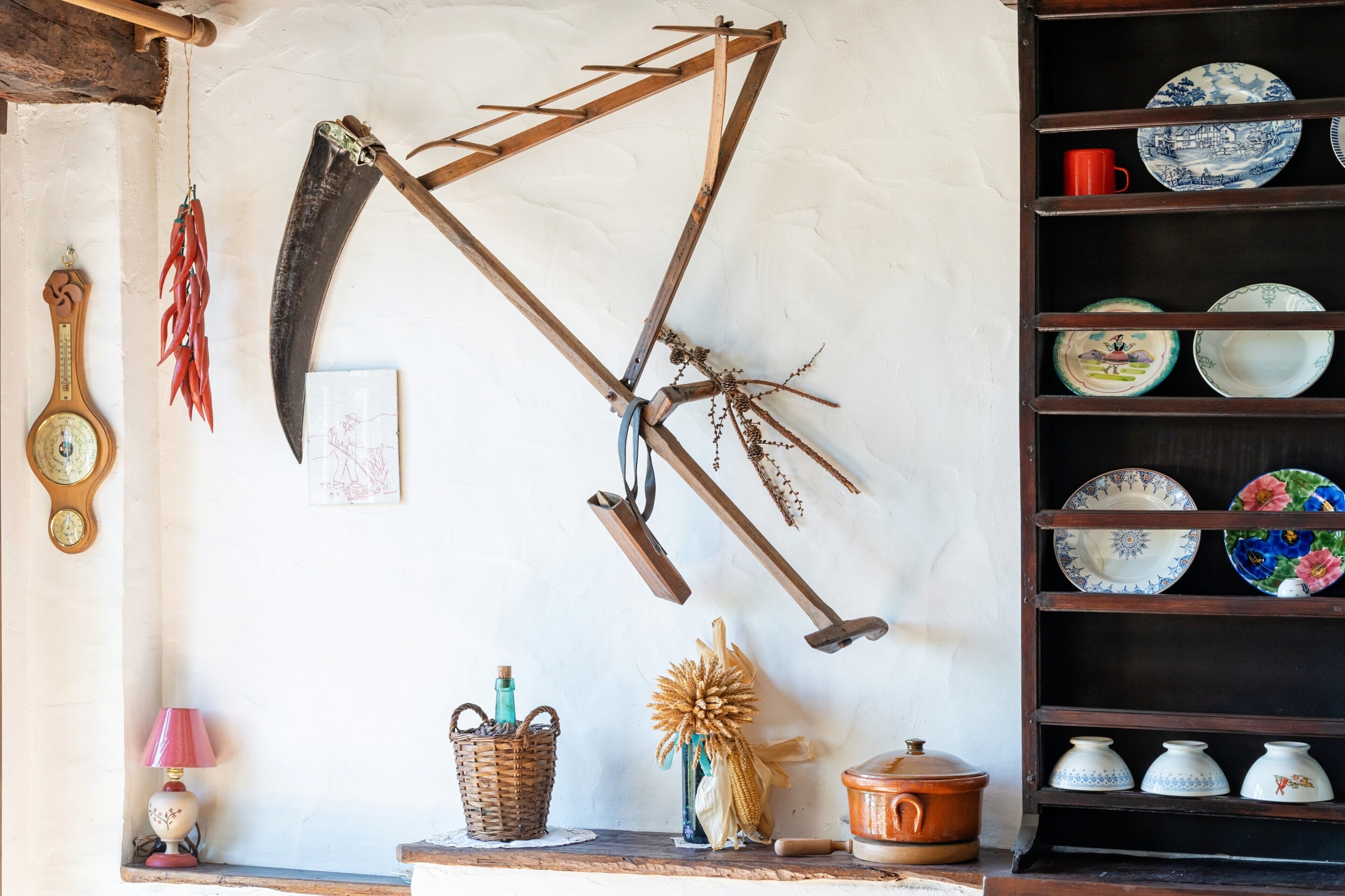 Un interior rústico con una estantería de madera con vajilla, una guadaña colgada en la pared y un reloj tradicional.