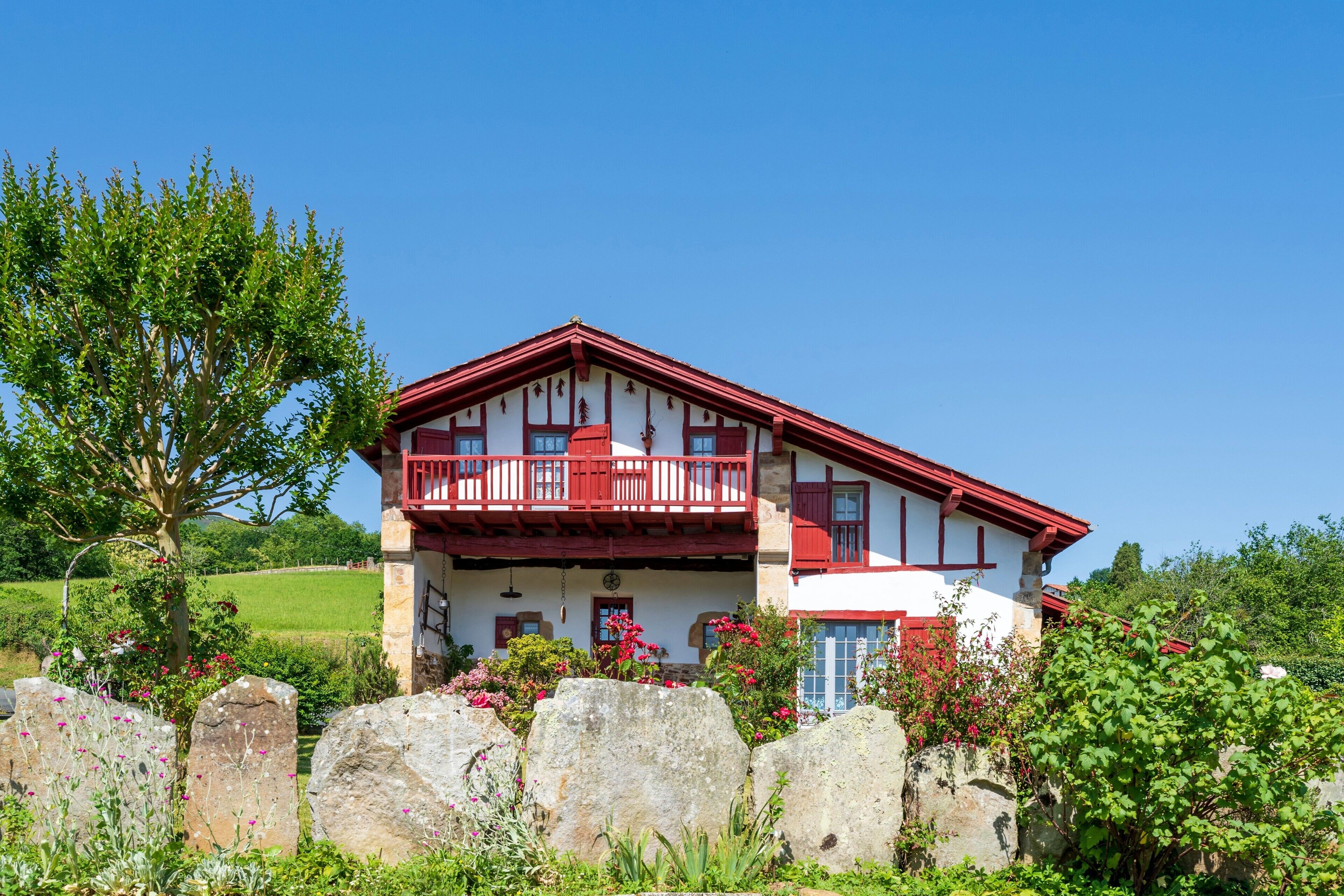 A two-story house with a balcony, surrounded by greenery and large rocks.