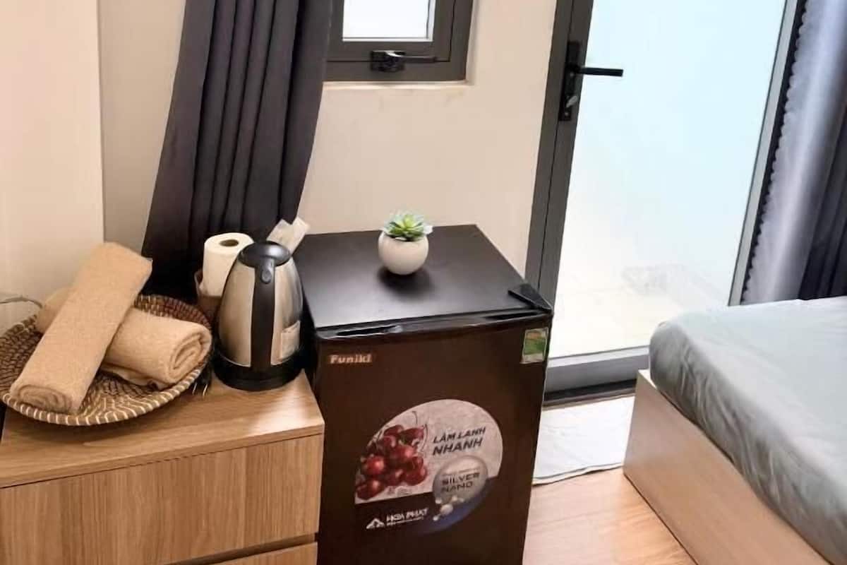 Ein Hotelzimmer mit einem Bett, einem kleinen Tisch mit einem Wasserkocher, einem zusammengerollten Handtuch, einer mit 'Funik' beschrifteten Box und einem Fenster mit Vorhängen.