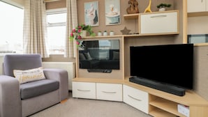 A living room with a grey sofa, a flat-screen TV, a wooden entertainment unit, and a window with curtains. - 21 Dunlin Dell (Bridlington)