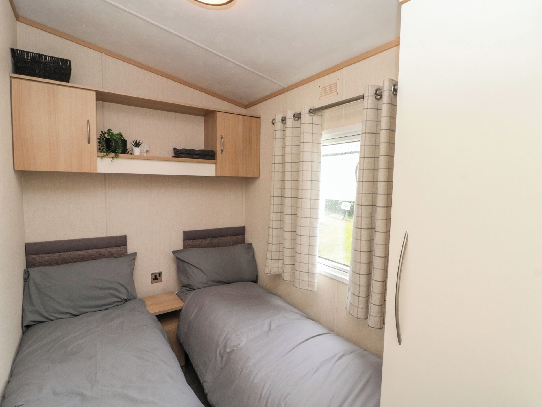 Un dormitorio pequeño con dos camas, una estantería con plantas y una ventana con cortinas.