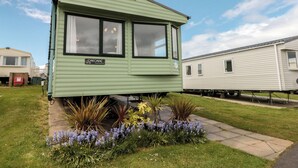 A green mobile home with a small porch, a nameplate, and a garden with various plants. - 21 Dunlin Dell (Bridlington)