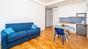 Un salon moderne avec un canapé et une table à manger, une cuisine avec des meubles blancs et un four à micro-ondes, et un parquet en bois disposé en chevrons.