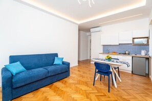 Ein modernes Wohnzimmer mit einer Couch und einem Esstisch, eine Küche mit weißen Schränken und einer Mikrowelle, sowie ein Holzboden mit Zickzackmuster.