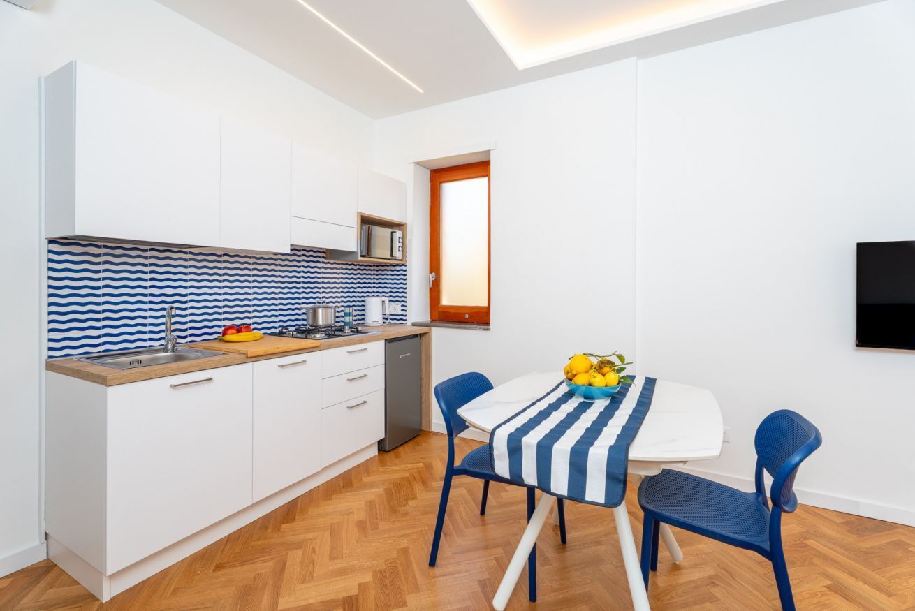 Cocina compacta con armarios blancos, un microondas y una encimera de madera. Hay una mesa de comedor con mantel a rayas azul y blanco y dos sillas azules.