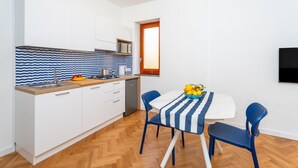 Une cuisine compacte avec des meubles blancs, un four à micro-ondes et un plan de travail en bois. On y trouve une table à manger recouverte d’une nappe à rayures bleues et blanches, ainsi que deux chaises bleues.