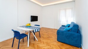 Un salon moderne avec un canapé bleu, une table à manger avec des chaises, un téléviseur à écran plat fixé au mur et un parquet en bois disposé en motif chevron.