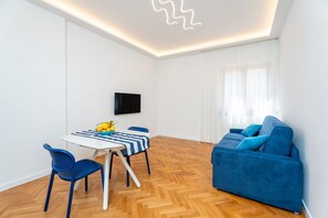 Ein modernes Wohnzimmer mit einem blauen Sofa, einem Esstisch mit Stühlen, einem an der Wand befestigten Flachbildfernseher und einem Holzboden mit Fischgrätmuster.