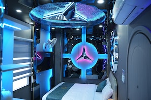 Une chambre moderne et compacte, dotée d’un lit, d’un luminaire rond et d’un ventilateur de plafond.