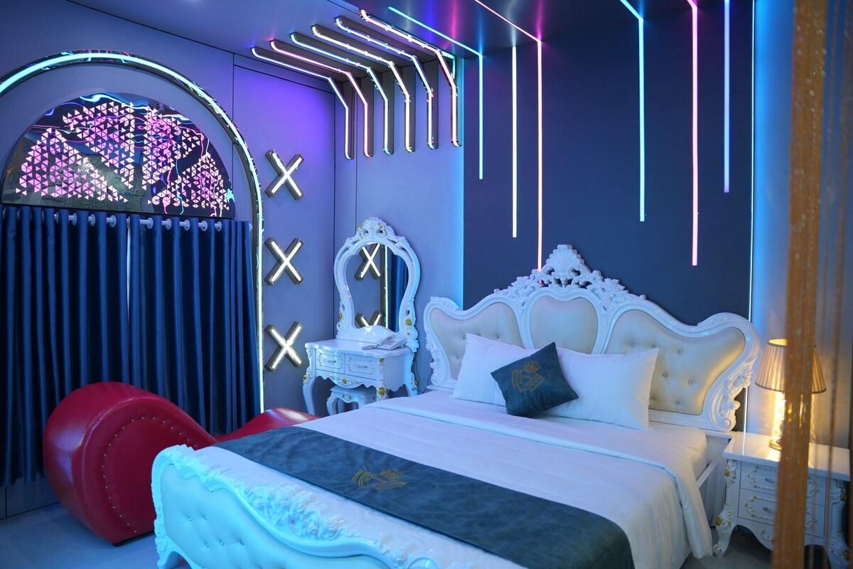 A bedroom with a white ornate bed, a red chair, a vanity with a mirror, and neon lights.