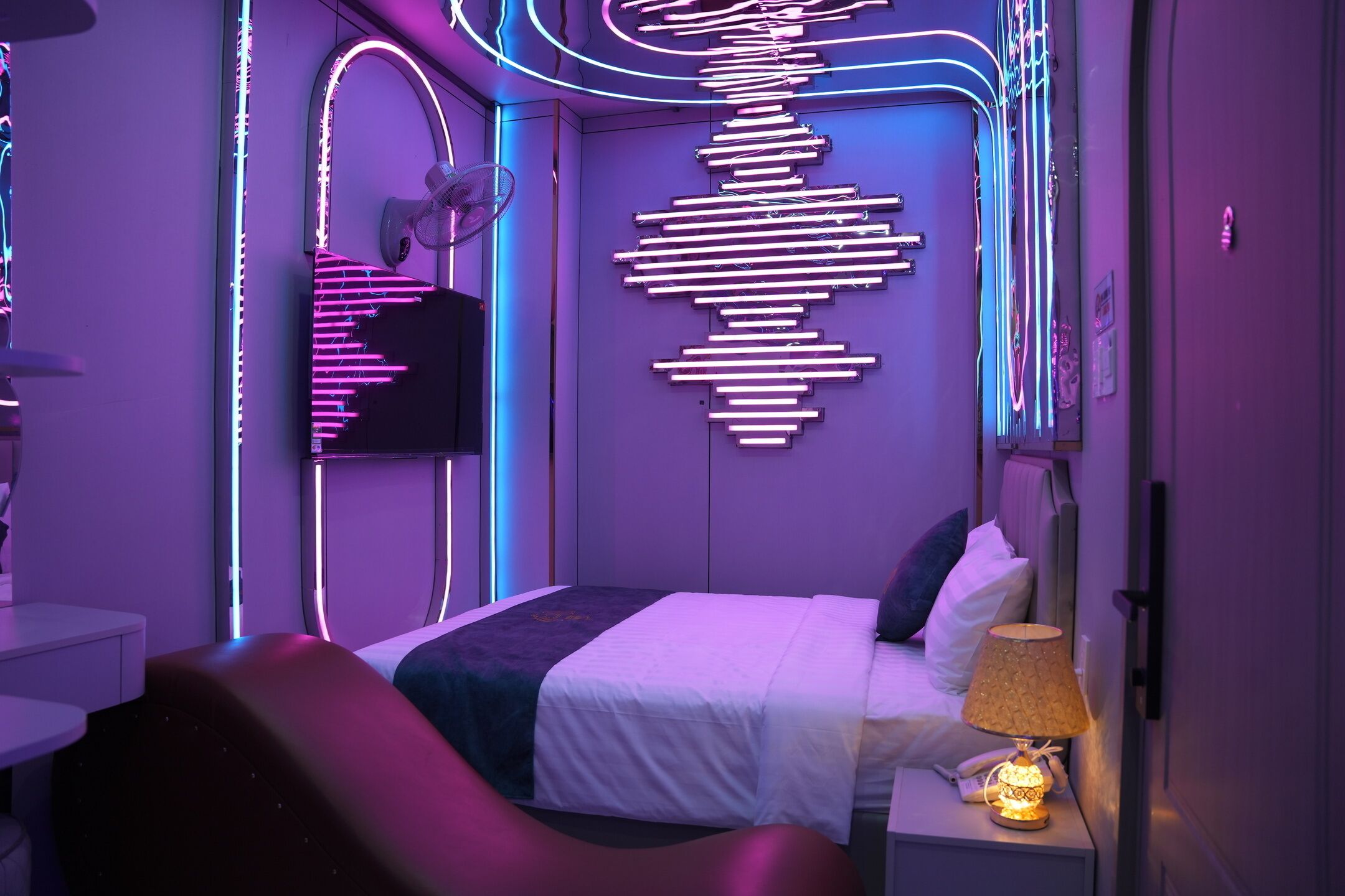 A modern hotel room with a bed, a bedside table with a lamp, a wall art installation, and a curved seating area.