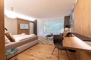A modern hotel room with a bed, a desk, a chair, a small table, a lamp, and a window with curtains.
