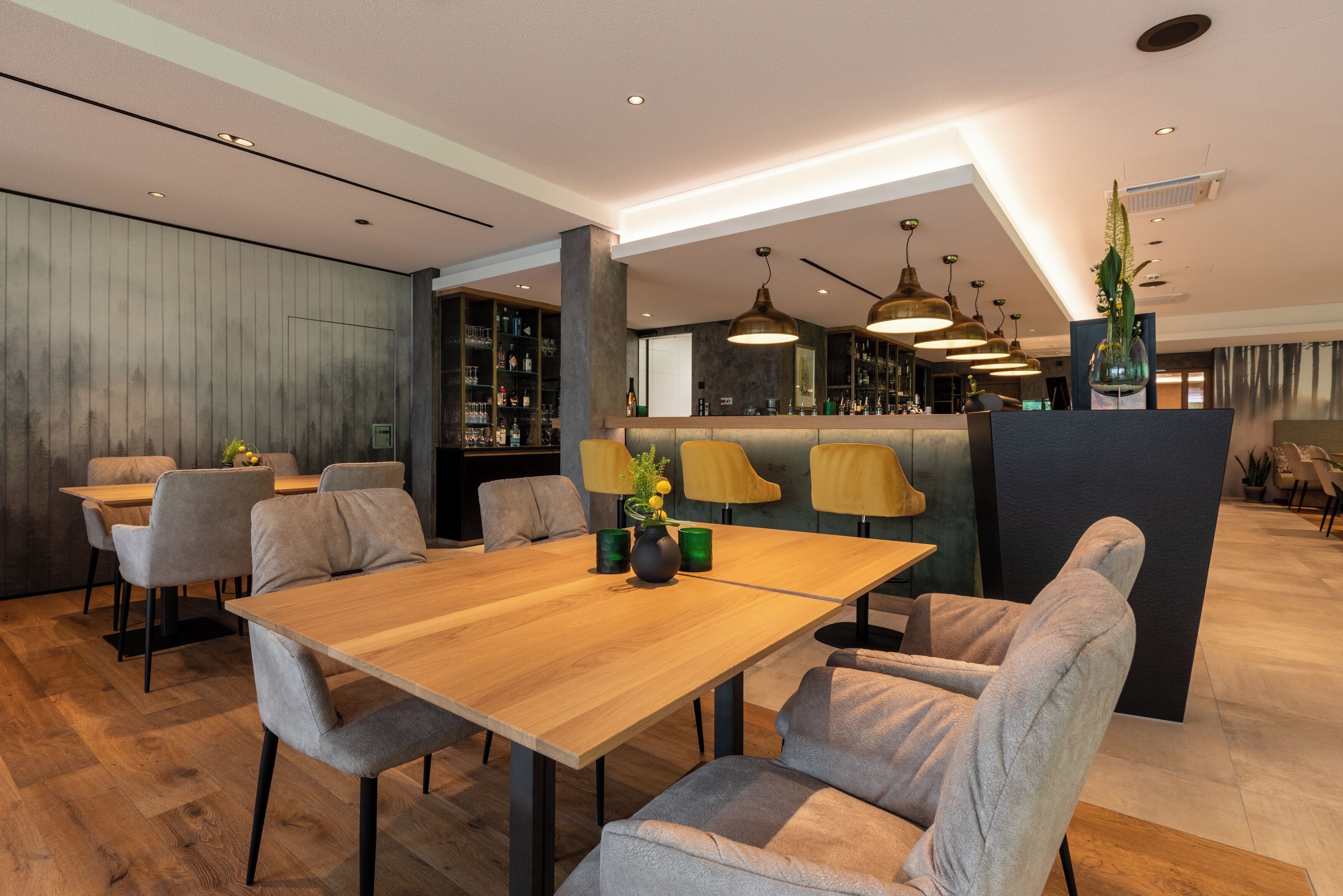 Un espace repas moderne avec une table en bois et des chaises à revêtement gris, un coin bar avec des étagères remplies de bouteilles et un éclairage suspendu.