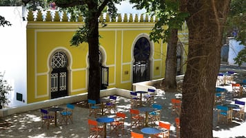 Un edificio amarillo con ventanas arqueadas y una puerta negra, rodeado de mesas al aire libre con sillas azules y anaranjadas.