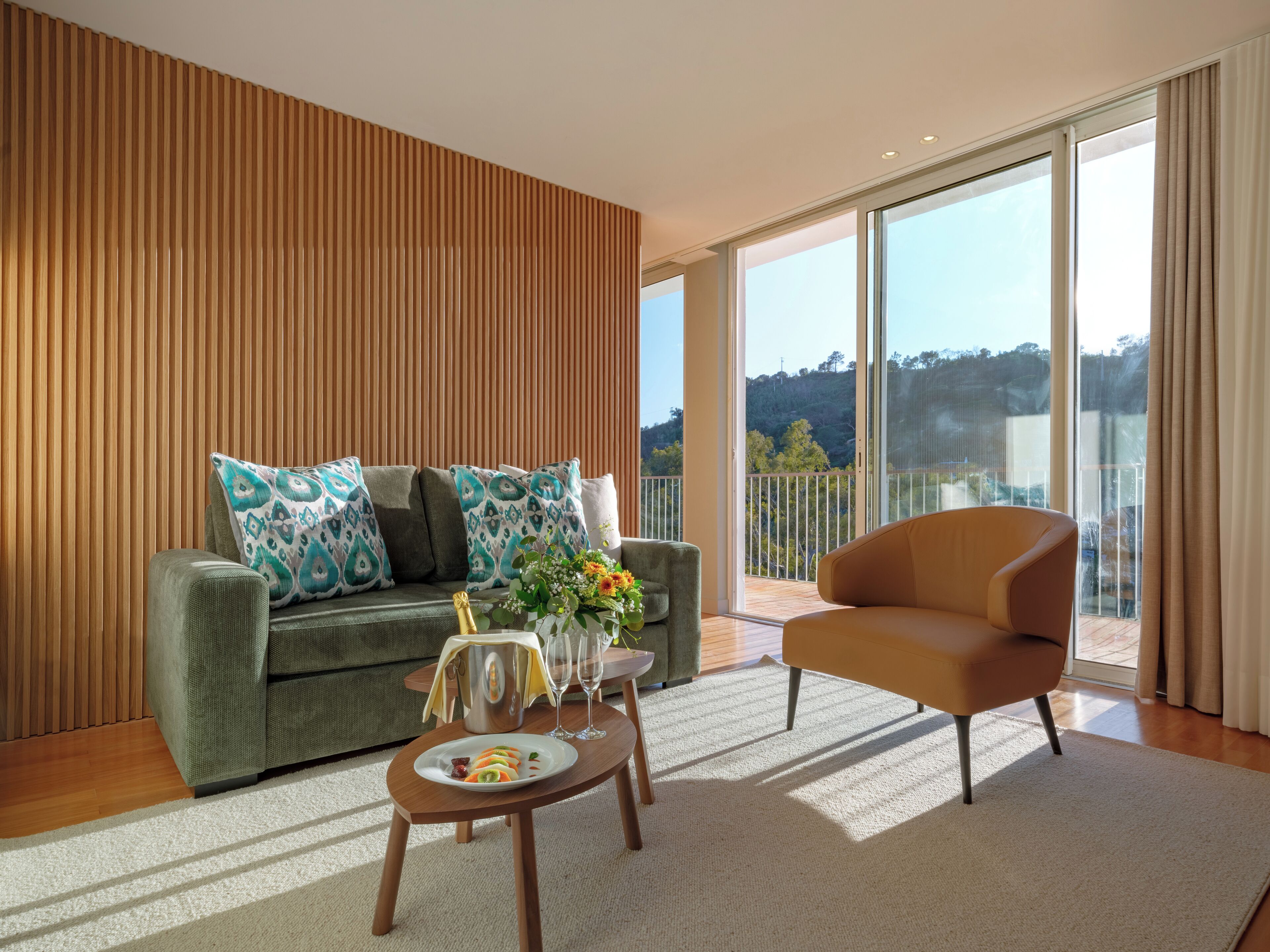 A modern living room with a green sofa, a wooden accent wall, a wooden coffee table with fruit, and a balcony with a view.