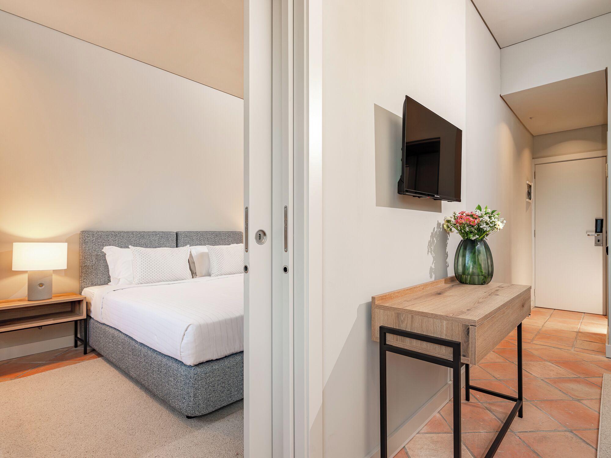 Habitación de hotel con cama, mesita de noche, lámpara, televisor montado en la pared y una pequeña mesa con un florero.