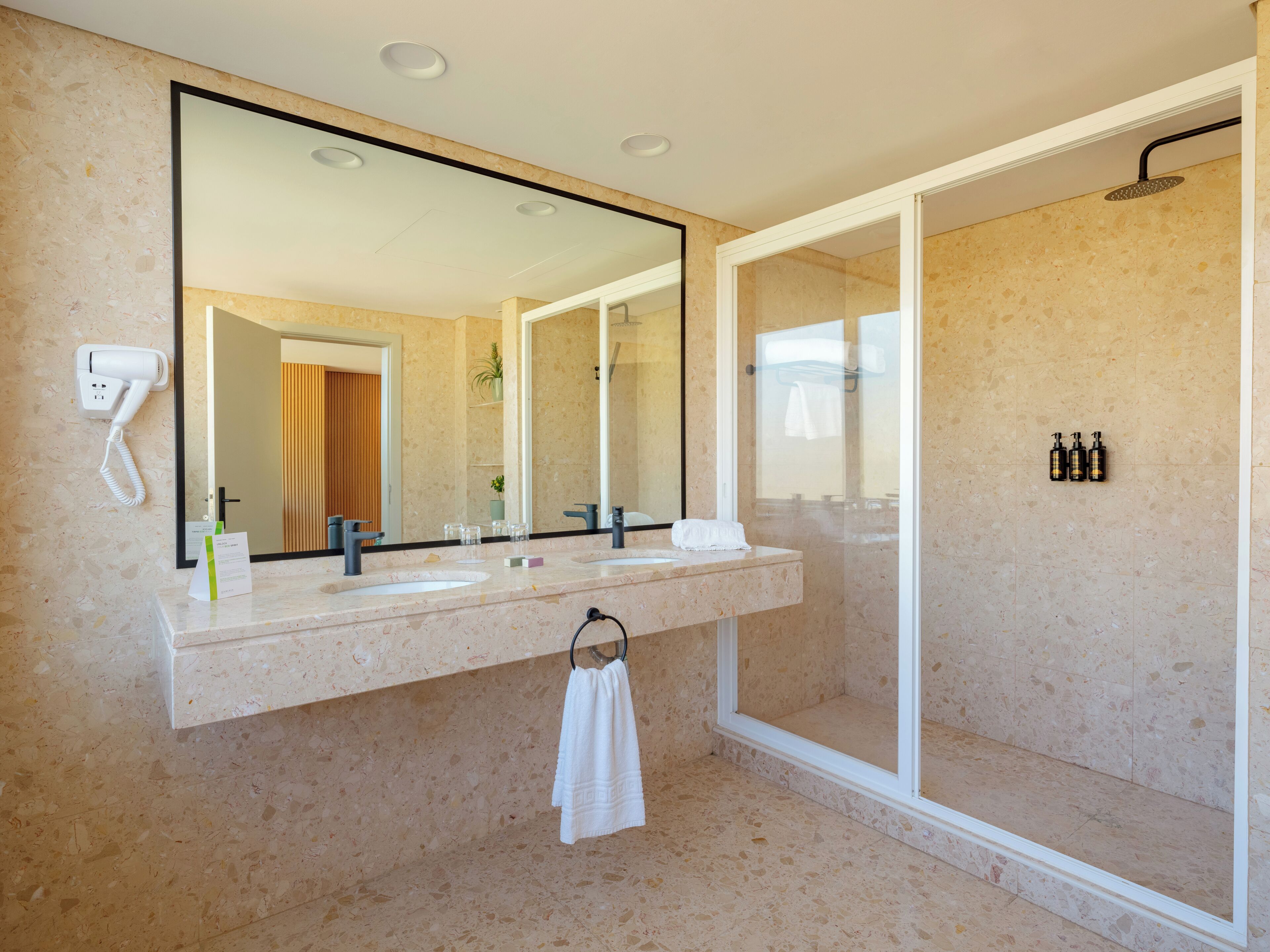 A modern bathroom with a large mirror, two sinks, a shower, and a hairdryer on the wall.