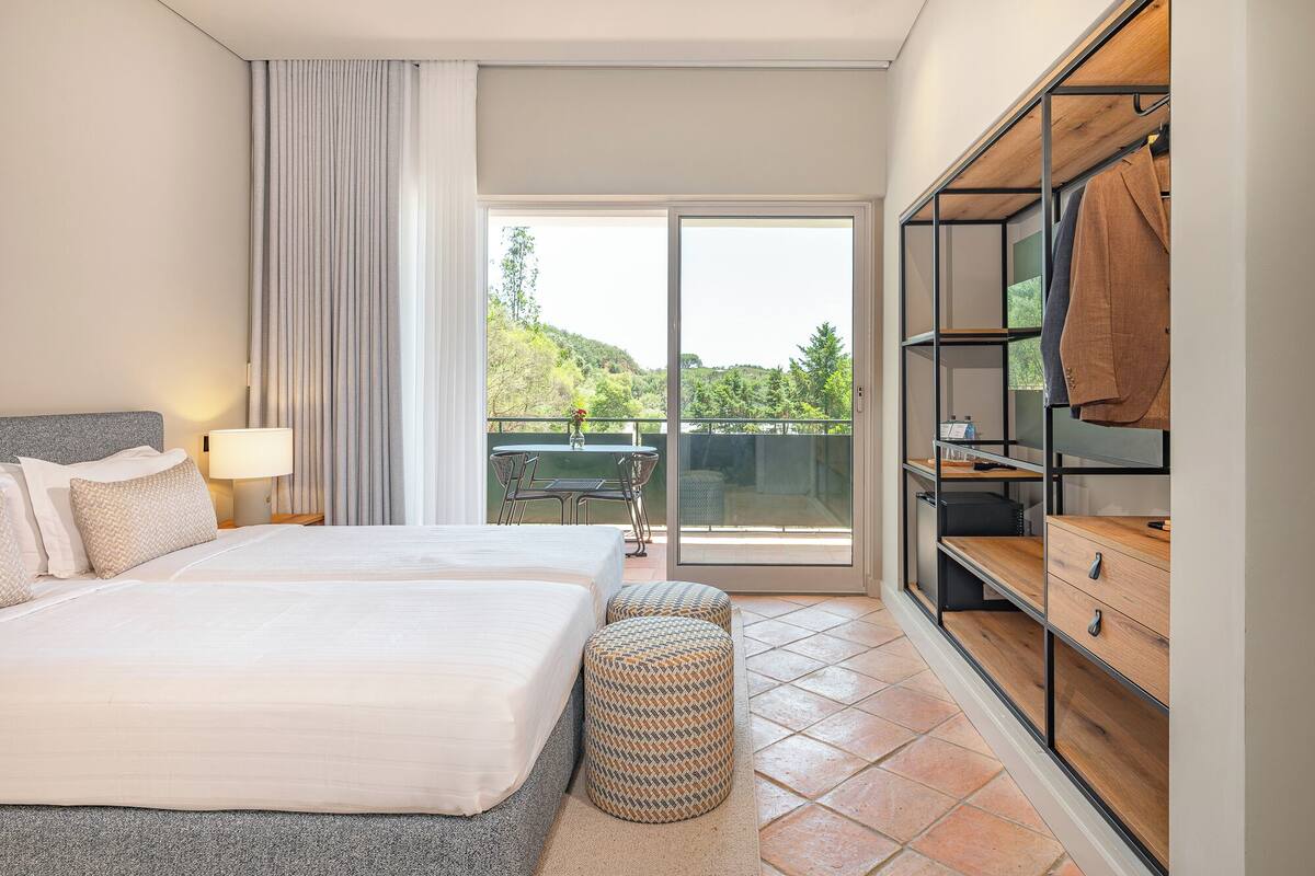 Habitación de hotel con cama, una mesita, una silla, armario y balcón con vistas a zonas verdes.