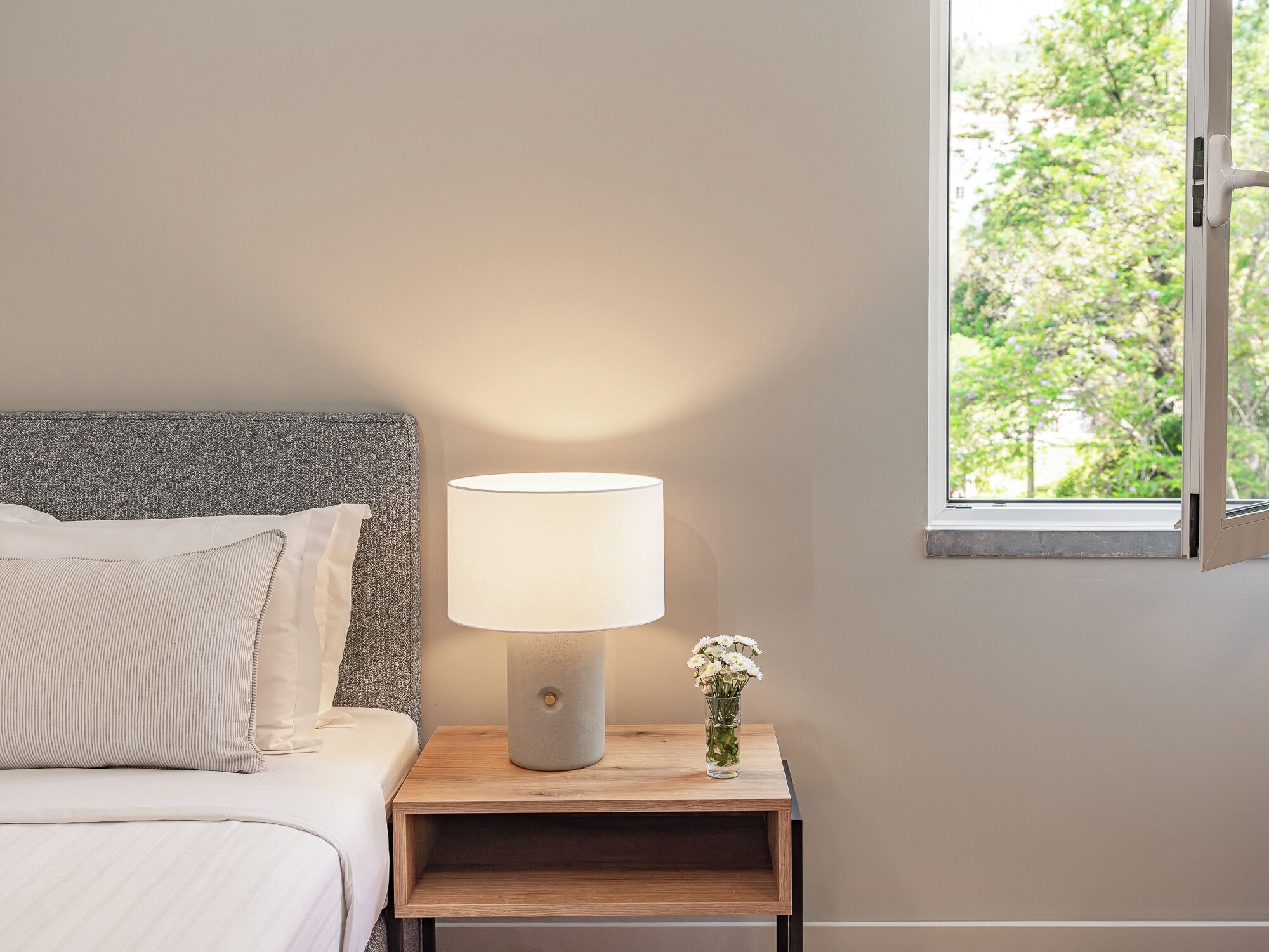 Ein Bett mit grauem Kopfteil, ein Nachttisch mit Lampe und ein Fenster mit Blick auf Bäume.