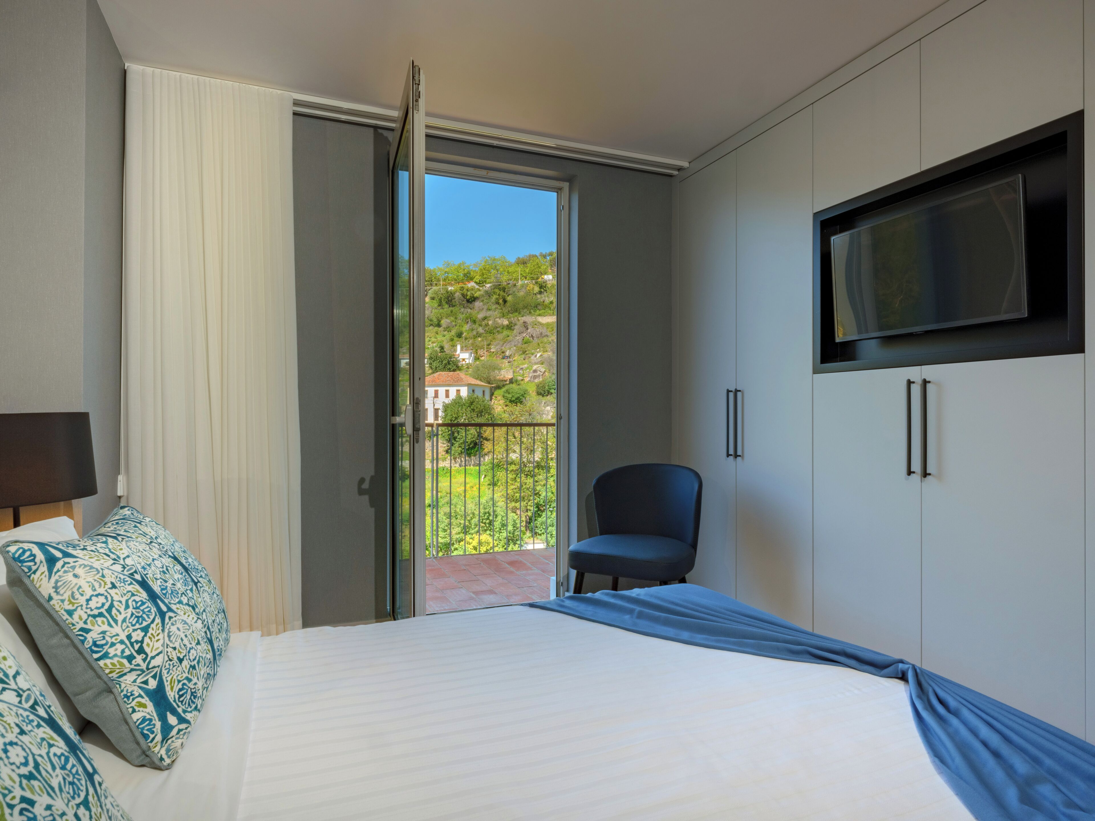 A hotel room with a bed, a chair, a TV, and a balcony with a view of houses and greenery.