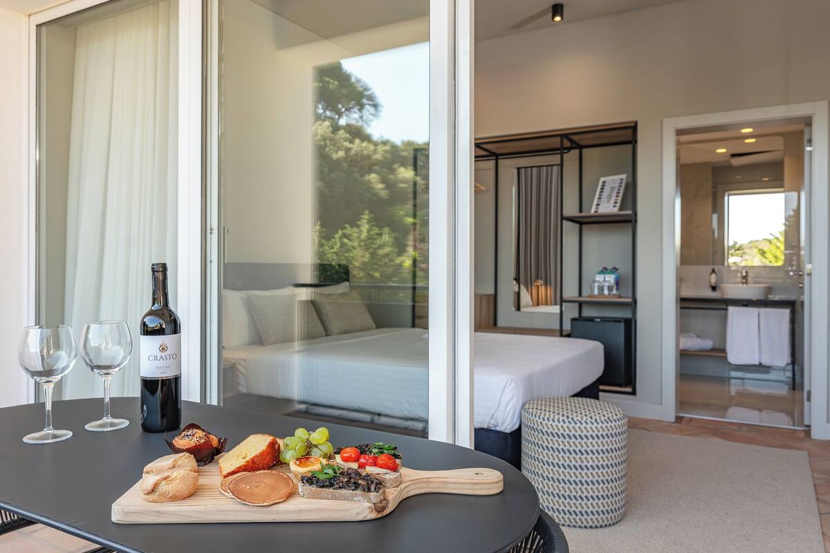 Una habitación de hotel moderna con cama, baño y comedor con mesa servida con comida y vino.