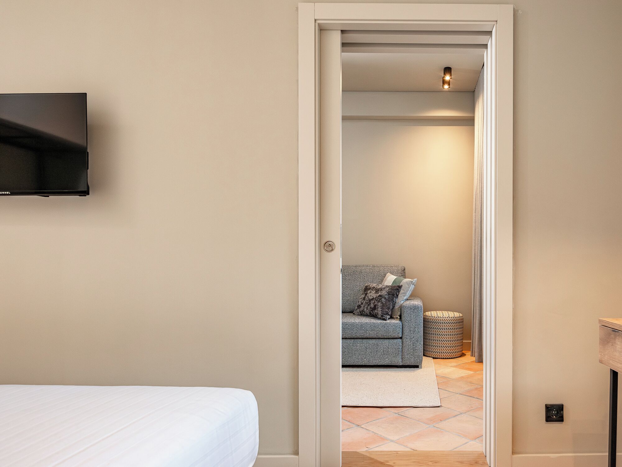 Une chambre à coucher avec un lit, un téléviseur fixé au mur et une porte donnant sur un espace de vie comprenant un canapé et une petite table ronde.