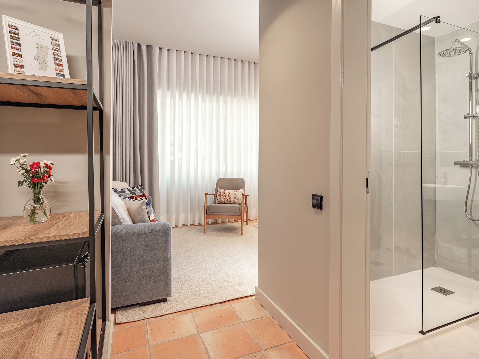 A modern bathroom with a glass shower enclosure, a wooden floor, and a seating area with a chair and cushions.