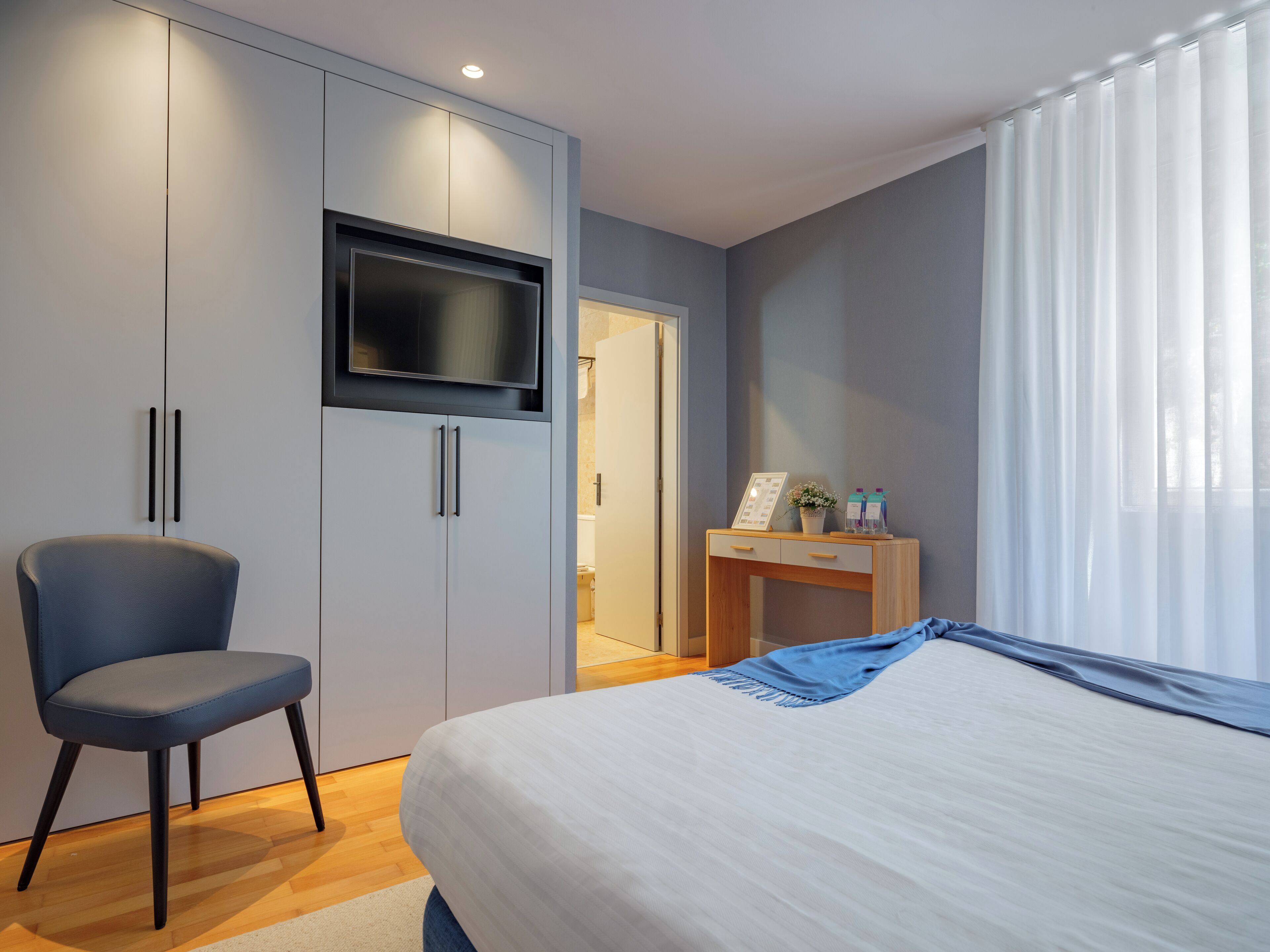 A bedroom with a bed, a chair, a TV mounted on a cabinet, and a desk with a plant and a picture frame.