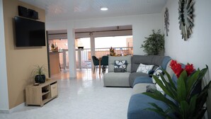 Un salon moderne avec un canapé gris, un téléviseur à écran plat, un meuble en bois et, au fond, un coin repas.