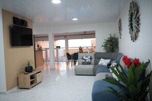 A modern living room with a grey sofa, a flat-screen TV, a wooden cabinet, and a dining area in the background.