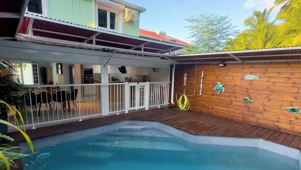 Une piscine avec une terrasse en bois, un patio couvert et une vue sur un bâtiment doté d’un toit rouge.