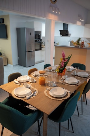 A dining area with a wooden table set for four, surrounded by teal chairs, with a kitchen in the background.