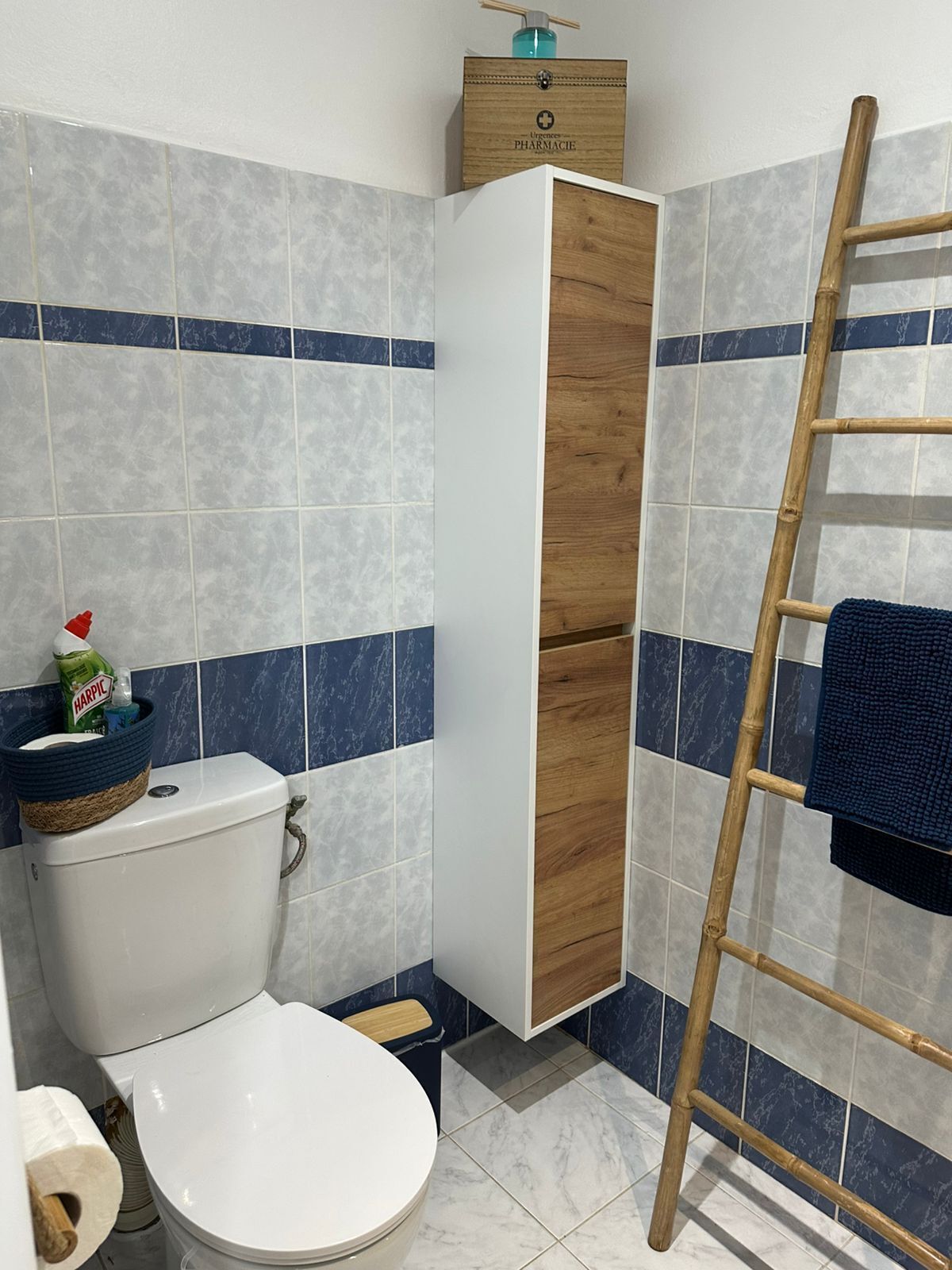 Um banheiro com vaso sanitário, armário de madeira, escada de bambu e toalha azul.