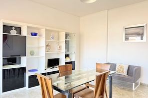 Een moderne woonkamer met een glazen eettafel, houten stoelen, een grijze bank en een wit tv-meubel met planken.