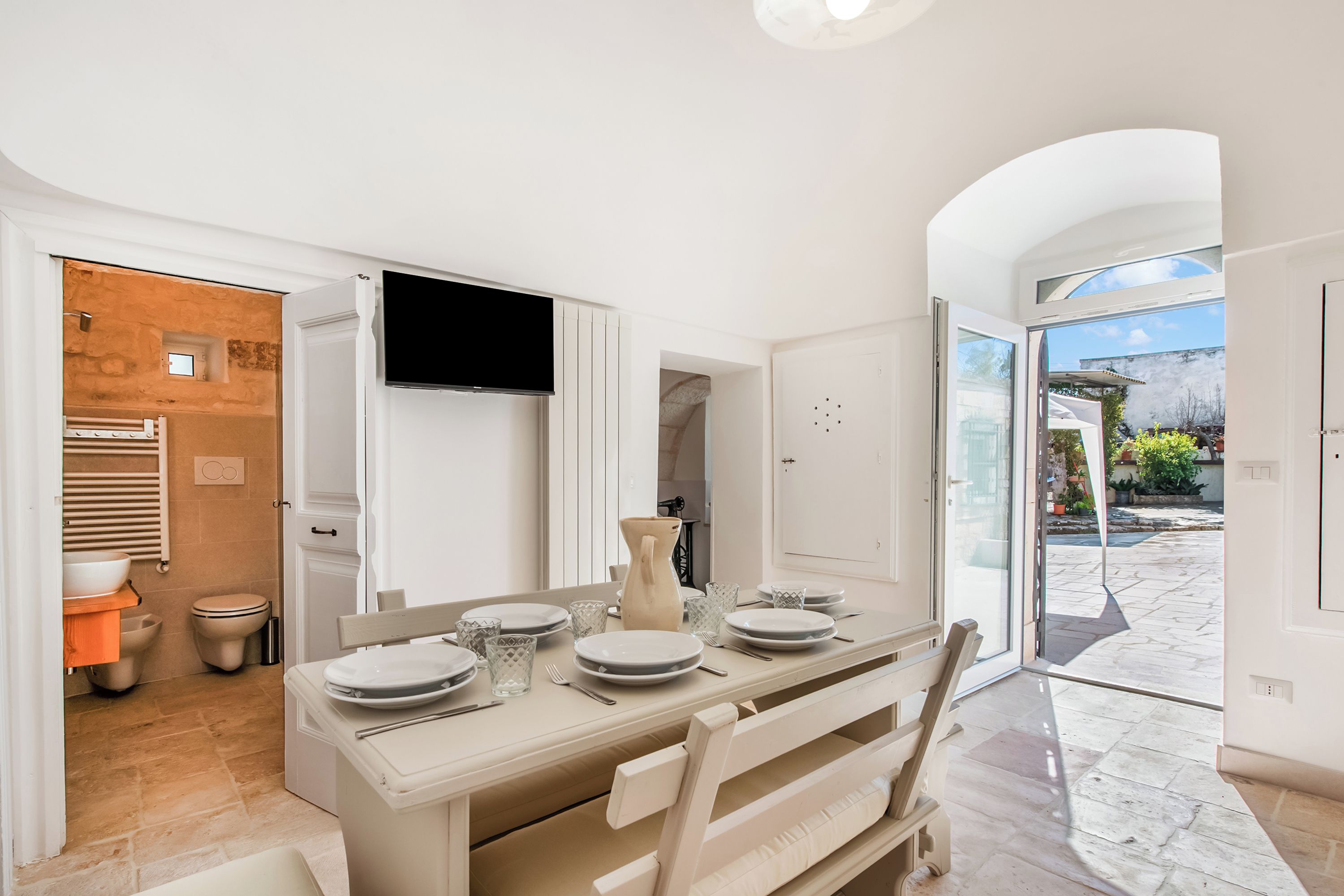 A modern kitchen with a flat-screen TV, a dining table set with plates and glasses, and a view of the outdoor patio.