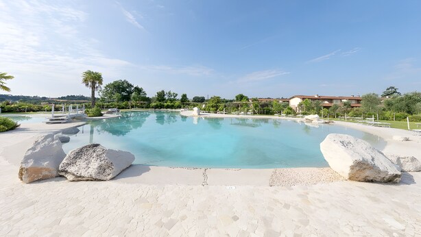 Una piscina blu cristallina con grandi rocce, circondata da una zona pavimentata e vegetazione.
