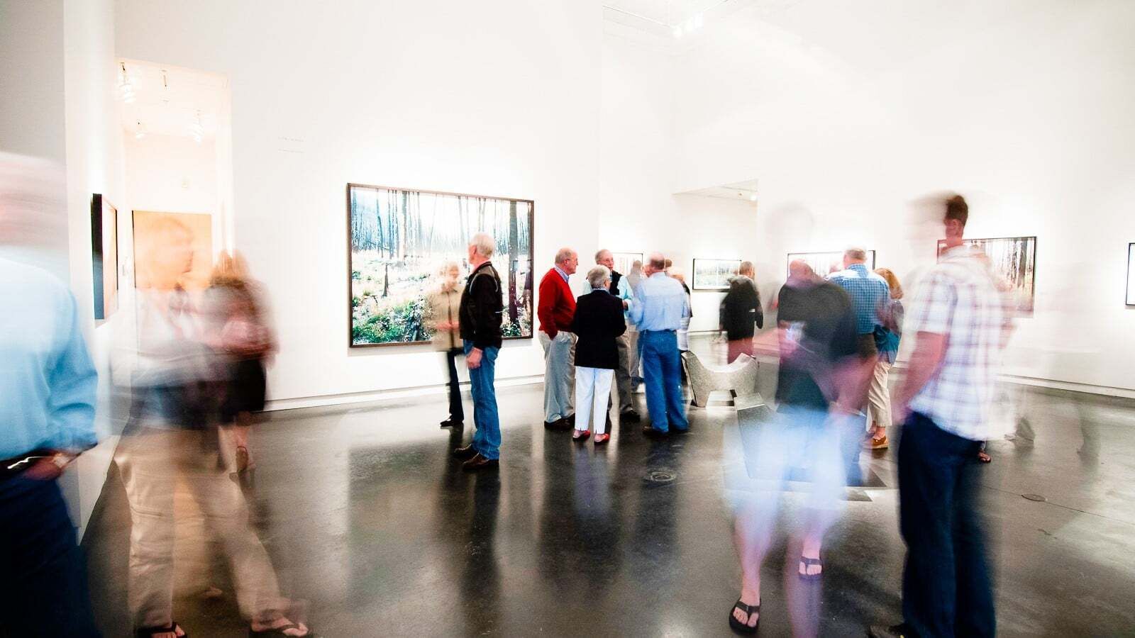 Una galería amplia con piso pulido, pinturas grandes en las paredes y un grupo de personas observando las obras de arte.