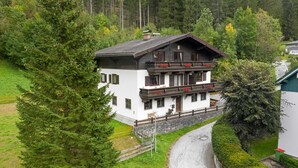 A two-story chalet with a balcony, surrounded by greenery and a forested backdrop. - Ferienhaus Höllbacher by we rent (Bad Gastein)