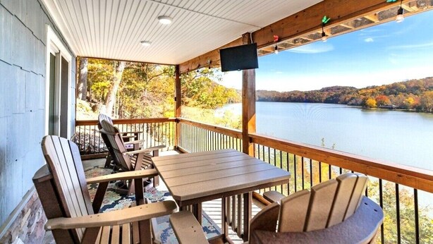 En träveranda med Adirondack-stolar och ett bord, med utsikt över en sjö omgiven av höstfärgat lövverk.