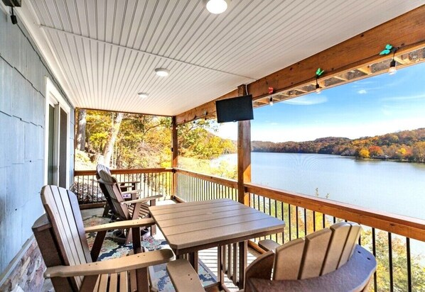 A wooden deck with Adirondack chairs and a table, overlooking a lake with autumn foliage. - Luxury Waterfront Cottage/Panoramic Views/Fully Renovated (Pulaski)