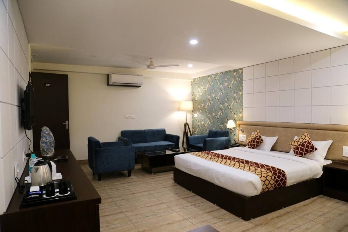 Habitación de hotel con una cama, dos sofás azules, un escritorio con un hervidor, una lámpara y un aire acondicionado instalado en la pared.