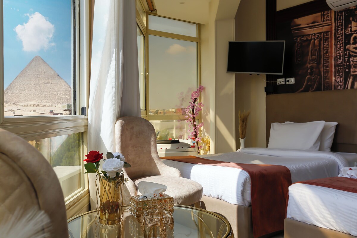 A hotel room with a bed, a chair, a glass table with a vase, and a view of a pyramid.