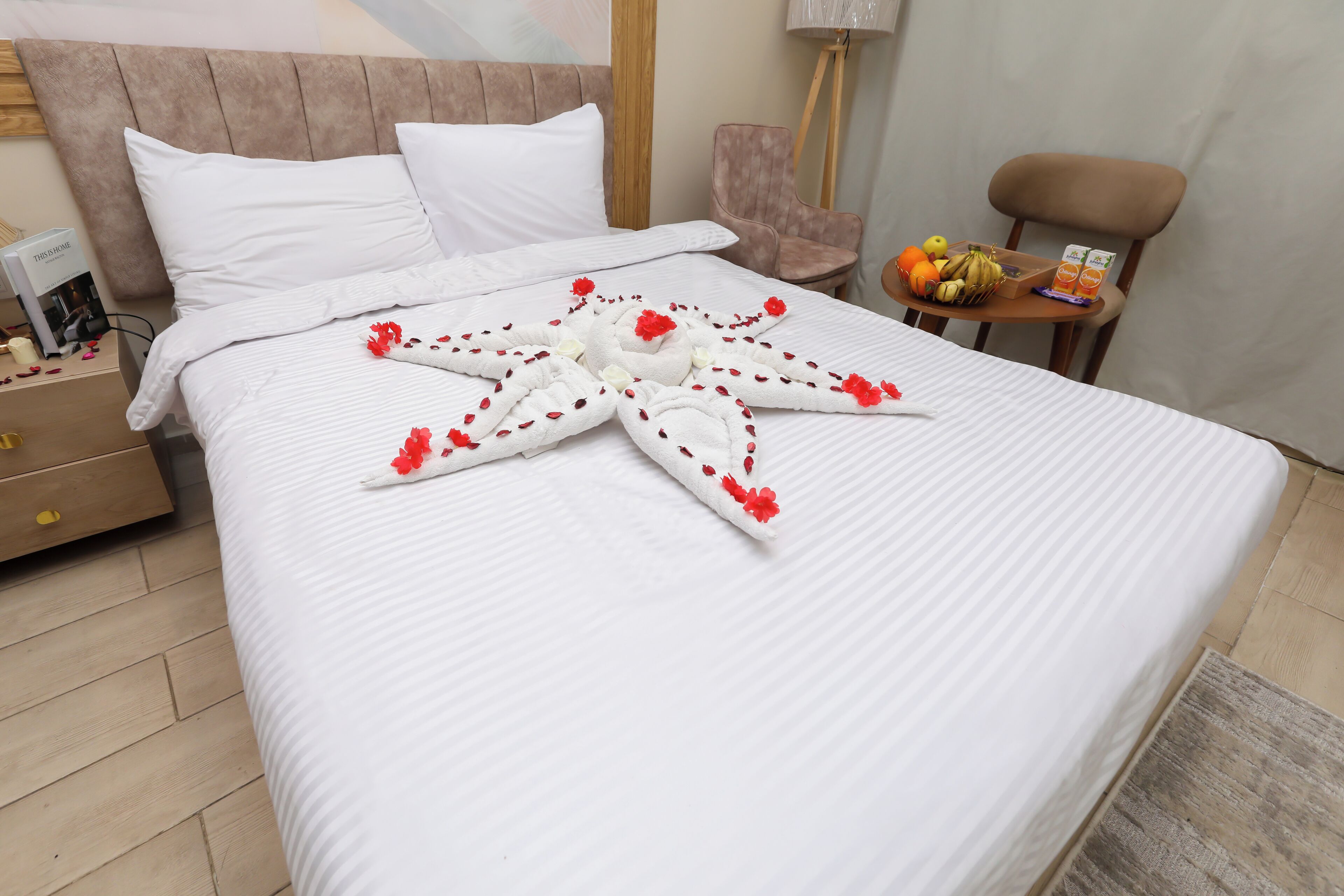 A hotel room with a neatly made bed, a bedside table with a lamp, a chair, and a small table with fruits and a juice box.