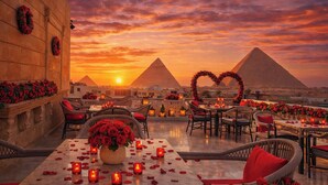 Un área de comedor al aire libre con mesas dispuestas para dos, rodeada de sillas de mimbre y decorada con rosas rojas y velas.