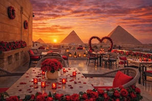 Un área de comedor al aire libre con mesas dispuestas para dos, rodeada de sillas de mimbre y decorada con rosas rojas y velas.