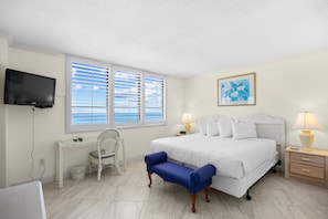 A bedroom with a bed, bedside lamps, a TV, a window with blinds, a chair, and a desk. - Arcadian 1 | 2 Bedroom Oceanfront Condo (Myrtle Beach)