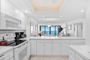 A modern kitchen with white cabinetry, a large island, a microwave, a kettle, and a coffee maker. - Arcadian 1 | 2 Bedroom Oceanfront Condo (Myrtle Beach)