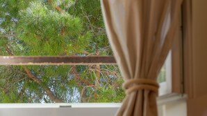 Una finestra con una tenda, un letto con un cuscino decorato e vista su alberi verdi all'esterno.