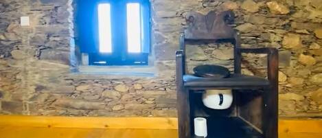 A room with a stone wall, a small window with vertical blinds, a wooden chair with a tray, and a wooden floor.